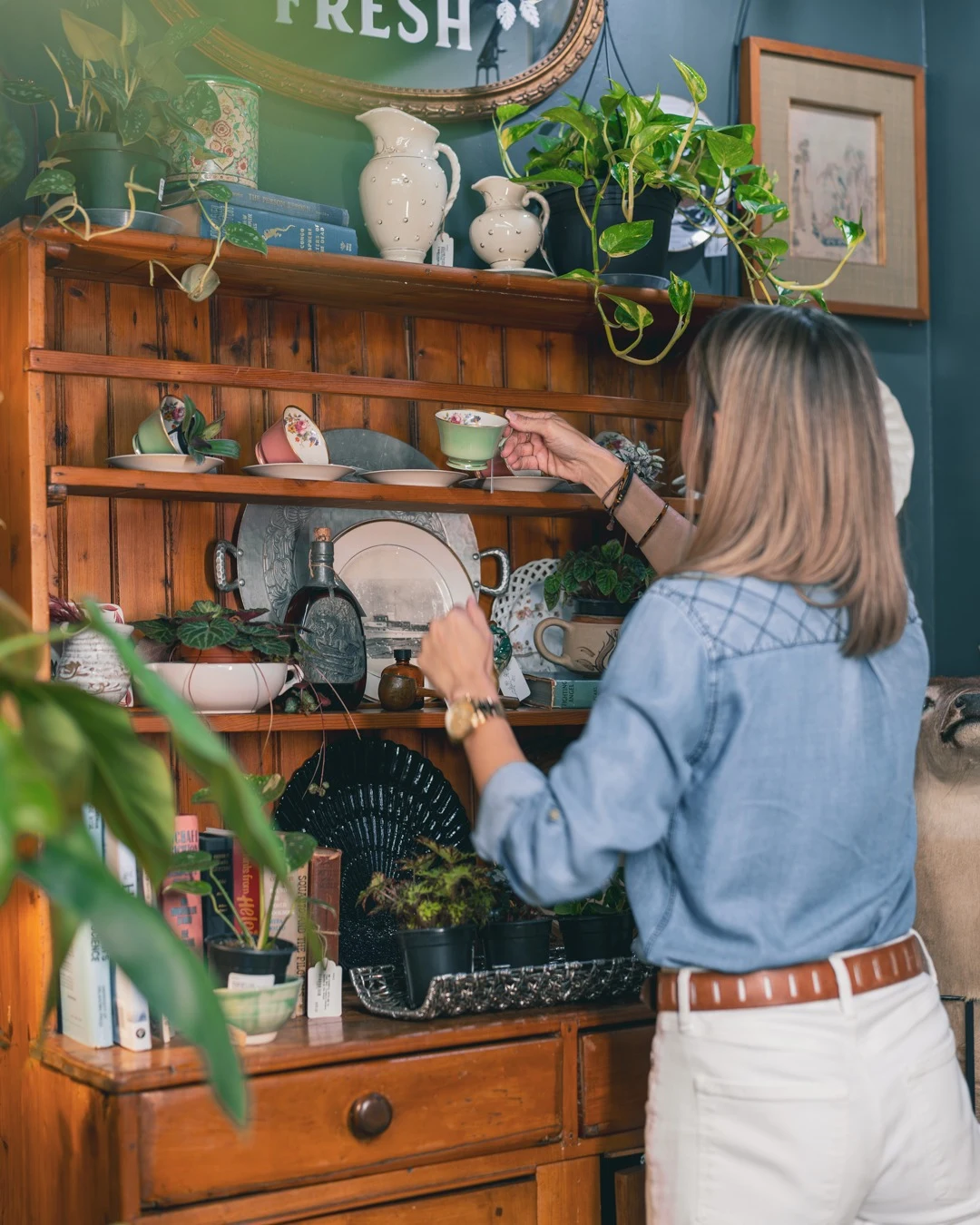 A shopper holding a vintage teacup beside a wooden hutch filled with plants and decorative finds